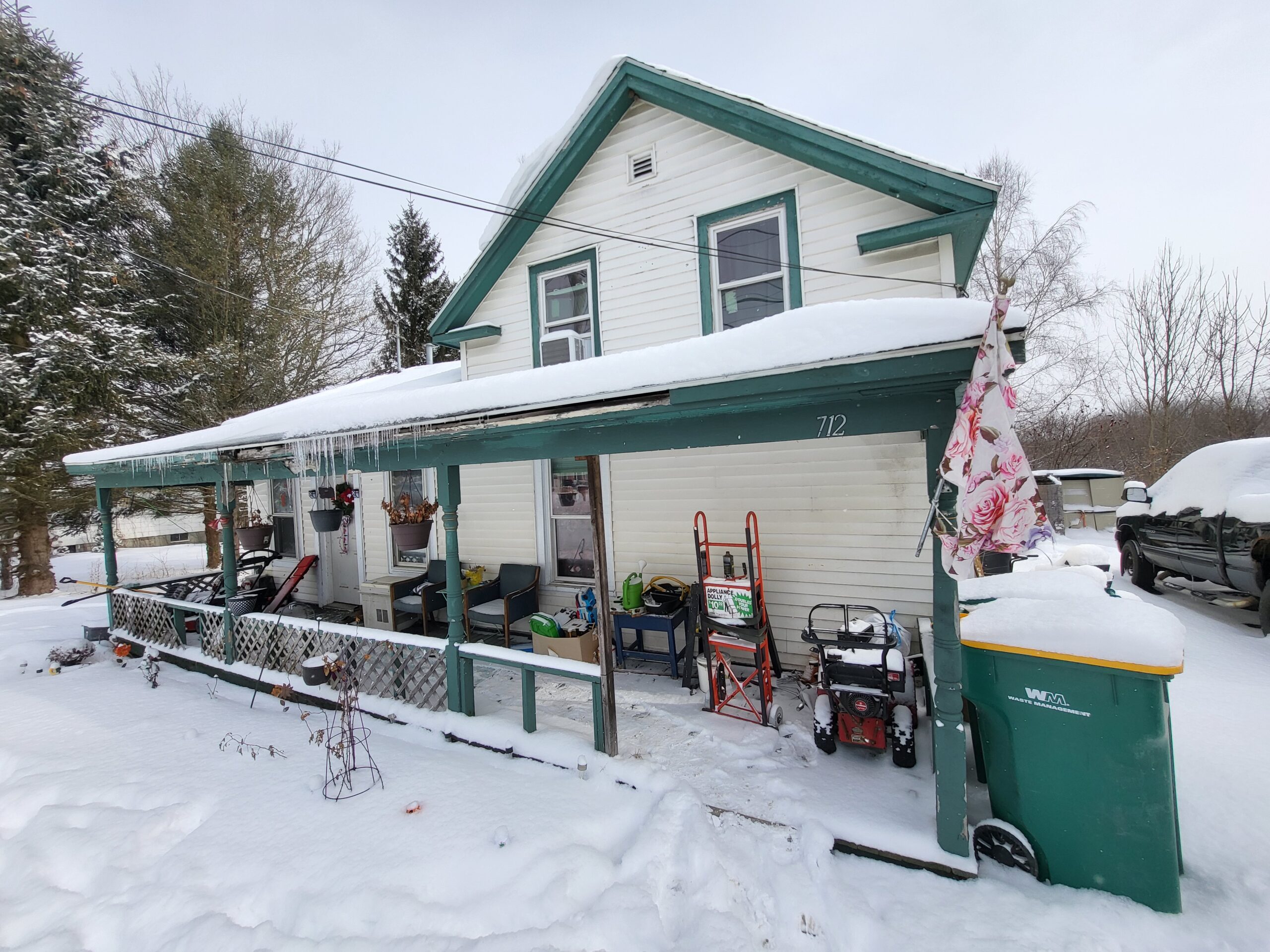 A white wooden house with a green trim is covered in snow. The porch has various items, including a snow blower, ladder, and outdoor furniture. A green waste bin is by the steps, and there's a snow-covered yard with trees in the background.