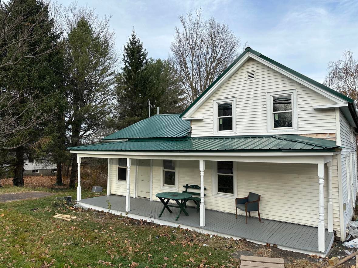 A white wooden house with a green trim is covered in snow. The porch has various items, including a snow blower, ladder, and outdoor furniture. A green waste bin is by the steps, and there's a snow-covered yard with trees in the background.